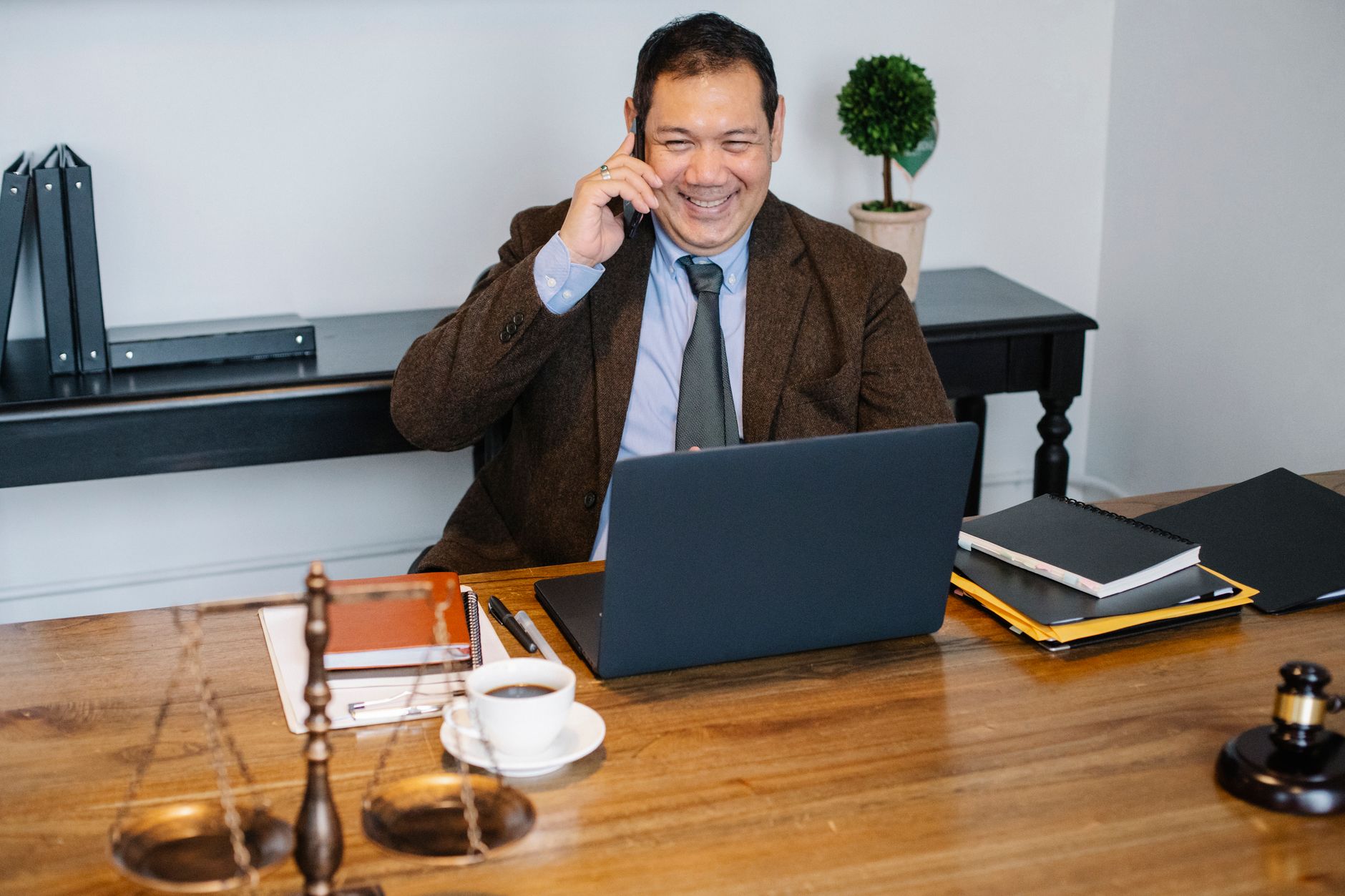 Advogado sorridente com terno formal conversando ao celular enquanto usa laptop em escritório moderno
